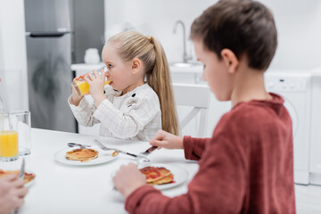Obraz na płótnie Canvas girl drinking orange juice near pancakes and blurred brother.