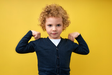 Look at my muscle, I'm strong. Portrait of confident little boy in T-shirt feeling powerful and self-confident, showing strength. indoor studio shot isolated on yellow studio background