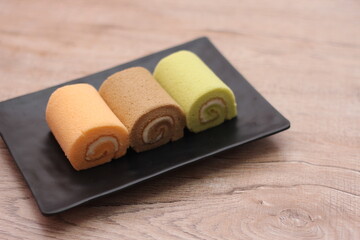3 Swiss rolls served in a black plate placed on a wooden table.