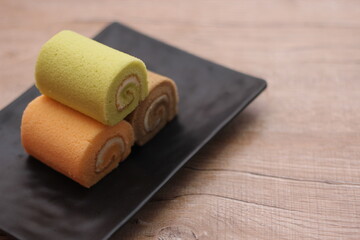 3 Swiss rolls served in a black plate placed on a wooden table.