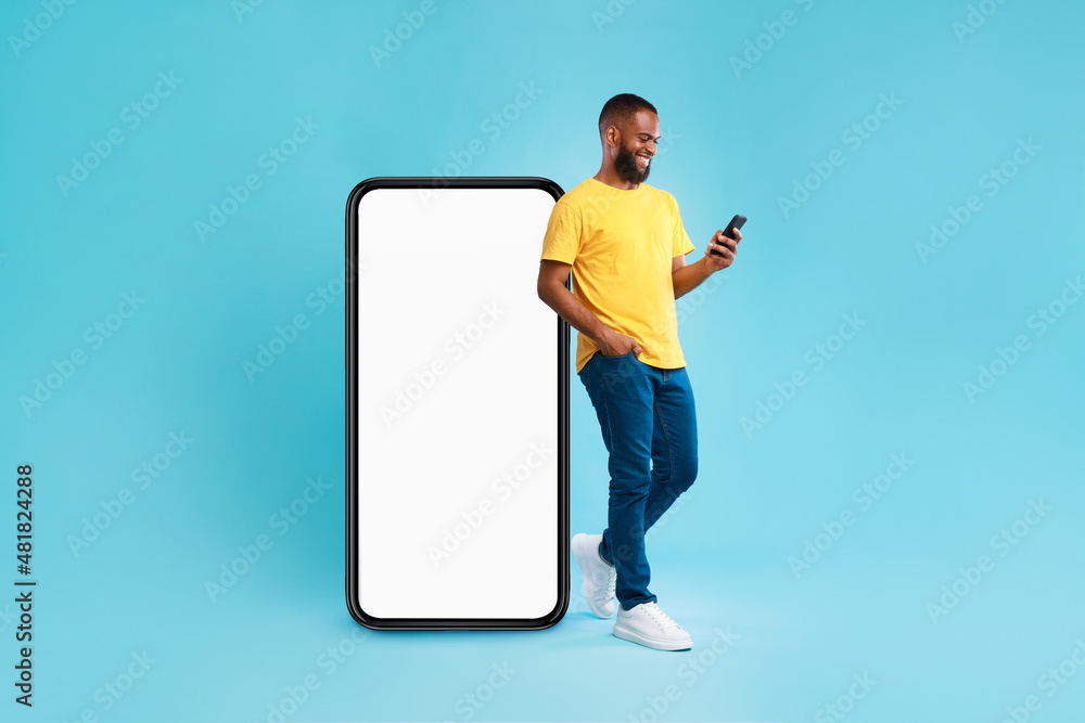 Aufkleber Handsome young Afro man using mobile phone while standing near huge smartphone with empty screen, mockup