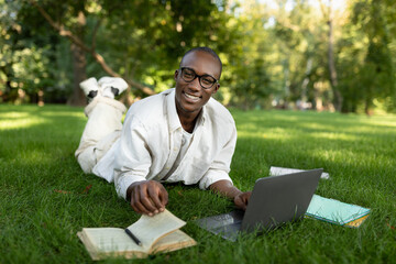 Happy african american student guy lying on grass at park, preparing for classes, using laptop and book outdoors