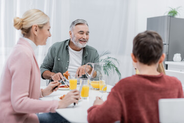 Fototapeta na wymiar cheerful senior man having breakfast with blurred grandchildren and wife in kitchen.