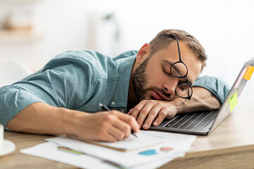 Exhausted millennial man sleeping on his office desk, next to laptop and documents, tired of overworking