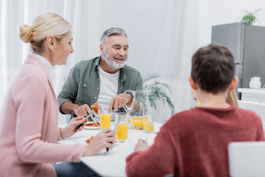 cheerful senior man having breakfast with blurred grandchildren and wife in kitchen.