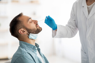 Coronavirus testing procedure. Doctor making nasal PCR test for millennial guy, using swab stick at...