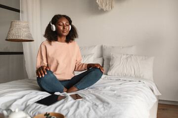 Black Woman Meditating Closing Eyes Wearing Headphones Relaxing In Bedroom