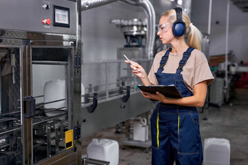 skilled engineer lady rate the quality of pesticides production in factory, take notes and think. professional female industrier holding folde-tablet and pen in hands, wearing uniform and headset