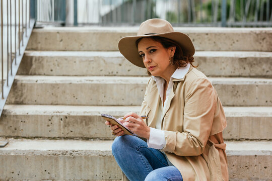 Woman taking notes and writing ideas in a notepad while sitting on the stairs in the street.