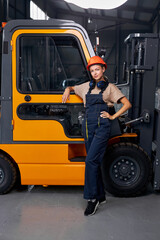 Attractive female standing next to forklift truck driver in an industrial area in factory, looking at camera confidently, dressed in uniform. good job. female engineer at work place