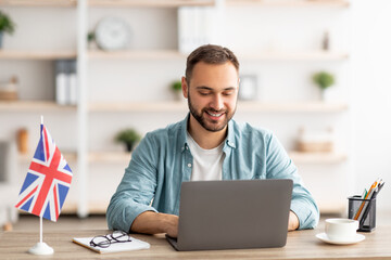 Happy young guy with flag of Great Britain working at desk with laptop in home office
