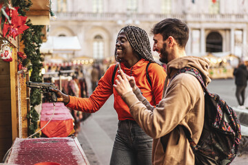 Couple of biracial young people tourists at the patronal feast they shoot the dartboard with the compressed air gun - the black girl with dreadlocks takes aim with the gun