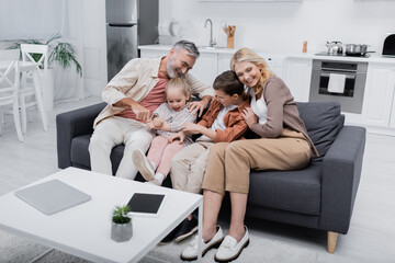 joyful couple hugging grandchildren near laptop and digital tablet with blank screen on table.