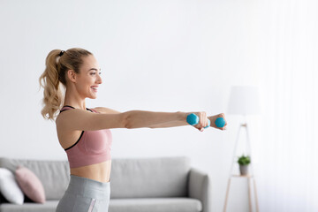 Fototapeta na wymiar Happy caucasian pretty young blonde lady lifting dumbbells at home in domestic gym, side view