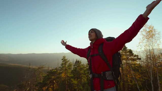 slow motion portrait of happy carefree girl hiker with backpack relaxing and enjoying sunset view raising arms at beautiful landscape in mountains, standing against blue sky at sunset. selective focus