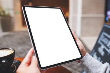 Mockup image of a woman holding digital tablet with blank white desktop screen with coffee cups on the table