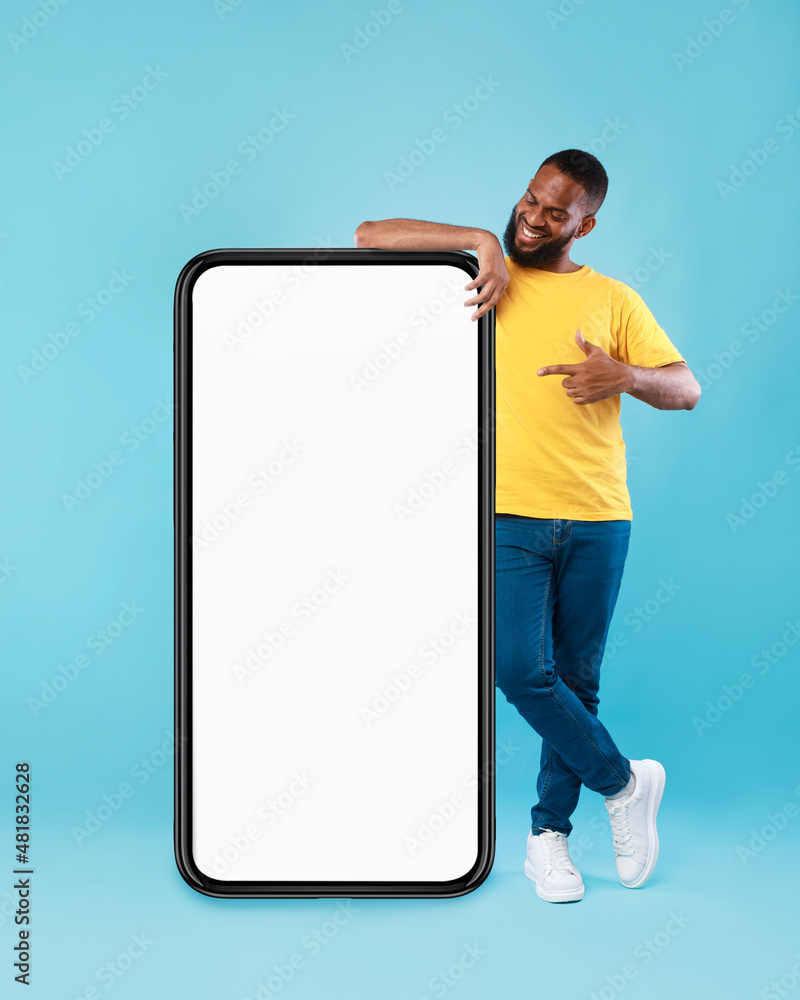 Poster cool young black guy pointing at empty smartphone over blue studio background, mobile application or