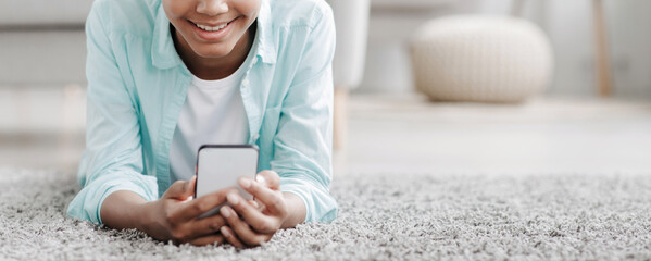 Smiling adolescent black girl watch online video, chatting in social networks lies on floor