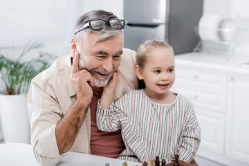 smiling girl touching face of happy grandpa playing chess in kitchen.