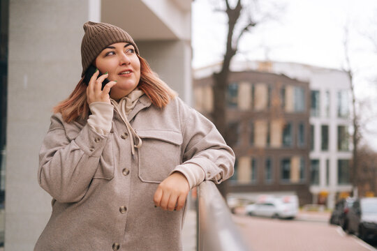 Portrait of attarctivve overweight woman in warm hat and jacket talking on mobile phone standing near railing at city street in cloudy autumn day. Frozen female having call on smartphone outdoors.