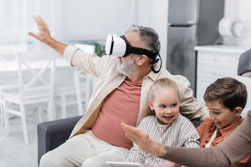 senior man in vr headset gesturing near grandchildren with digital tablet.