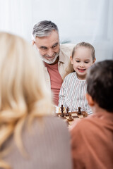 senior man smiling while playing chess with grandchildren and wife, blurred foreground.