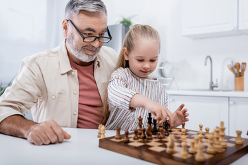 senior man in eyeglasses smiling near granddaughter playing chess.
