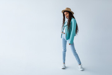 Full length photo of brunette girl with curly hairstyle in a mint jacket, blue jeans, white sneakers and a camel color hat. Cheerful African American Girl posing on light background. studio shot  