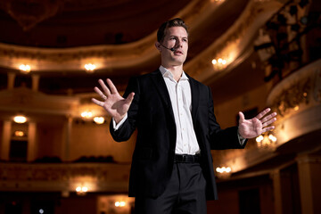 Attractive businessman standing on stage. Star, show, speech and presentation concept. Caucasian Handsome Male Presenter Speaking to Audience People in Conference Hall Auditorium, Gesturing.