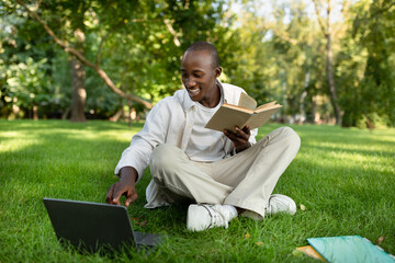 Preparing for exams outdoors. Happy black student guy studying with book and laptop computer, sitting in campus