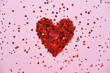 Heart made of shiny red small decorative hearts on a pink background strewn with sparkles. Flat lay, place for text. Festive minimal composition.