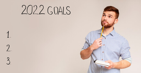 Thoughtful young man making 2022 goals list, dreaming, making new year resolutions on grey...