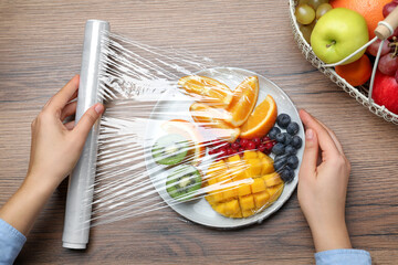 Woman putting plastic food wrap over plate of fresh fruits and berries at wooden table, top view