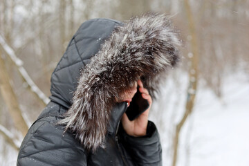 Woman in fur hood talking on mobile phone standing on winter street during snow. Cold weather,...