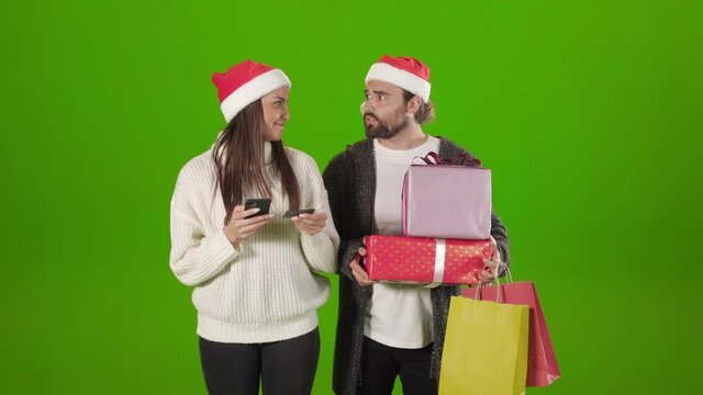 Young woman in santa hat using mobile and credit card for online shopping. Bearded man with christmas gifts and shopping bags in hands feeling sick because of prices.