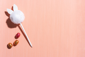 A white pen with a pompon on an orange background and chocolate eggs. The pen looks like a rabbit
