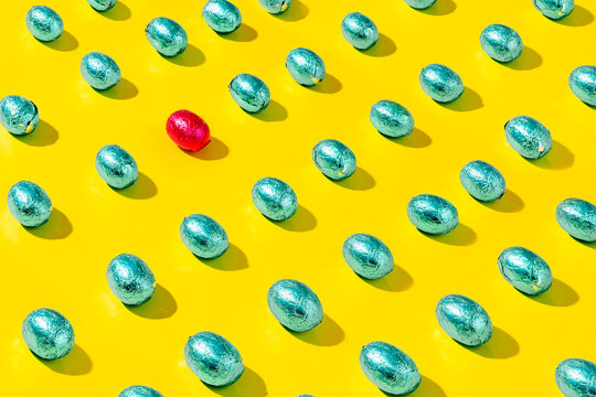 Pattern of green foil chocolate eggs on a yellow background. There is a red one which is different