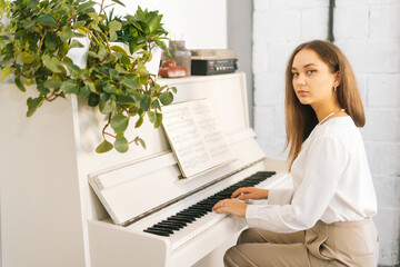 Portrait of serious pretty female pianist playing on white classical piano in light room, looking...