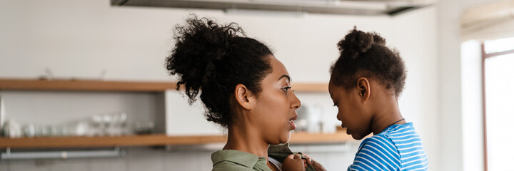 Black mid mother holding and talking with her daughter at home kitchen