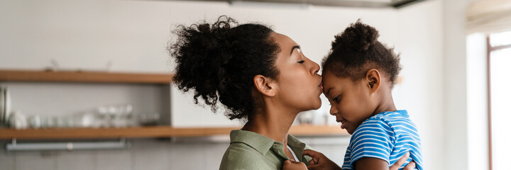 Black mother kissing and holding her daughter at home kitchen