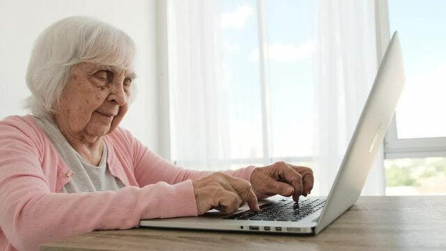 Senior woman typing on laptop keyboard and smiling. Elderly female person discovering modern technologies