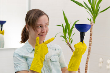 A young woman cleaner in yellow sanitary rubber gloves in a bright bathroom with a plunger to clear the blockage in her hands. Selective focus. Portrait