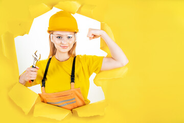 Young blond woman in uniform builder clothes, hardhat is holding metal wrench. Girl worker is...