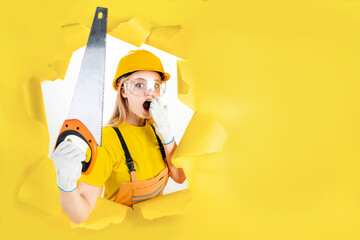 Energy fun handyman blond woman in protective helmet is holding manual saw in torn hole of yellow...