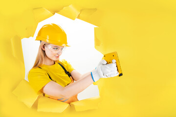 Serious blond woman worker is holding repair instrument, tool, accessory in torn hole of yellow...