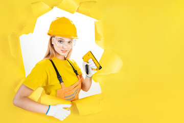Serious blond woman worker is holding repair instrument, tool, accessory in torn hole of yellow background. Female engineer in uniform and helmet is ready to renovate house flat. Hard work concept.