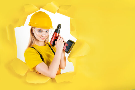Young employee worker woman in protective helmet is holding red screwdriver in torn hole of yellow background. Instruments, accessories, tools for renovation apartment room. Repair, build concept.