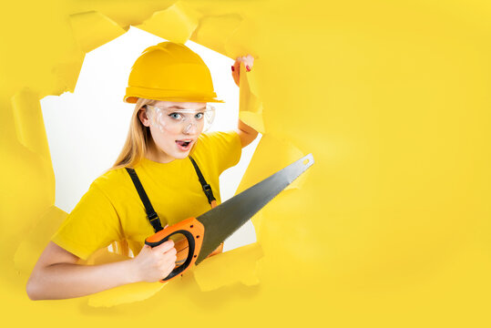 Energy fun handyman blond woman in protective helmet is holding manual saw in torn hole of yellow background. Female in male hard work. Renovation, repair, building concept. Emotional portrait.
