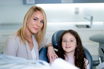 Portrait of a smiling little girl and her mother visiting a dentist. A little girl sitting in a dental chair and her mother standing near her.