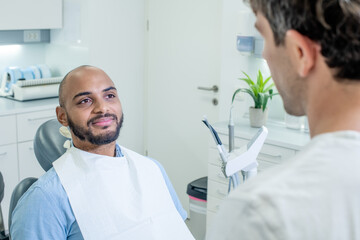 Fototapeta premium Over the shoulder shot of a male dentist talking reassuringly to his African American male patient, who smiling and looking relaxed while sitting in the dental chair.